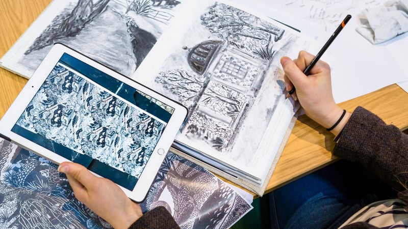 A student draws in a sketchbook using an ipad as reference