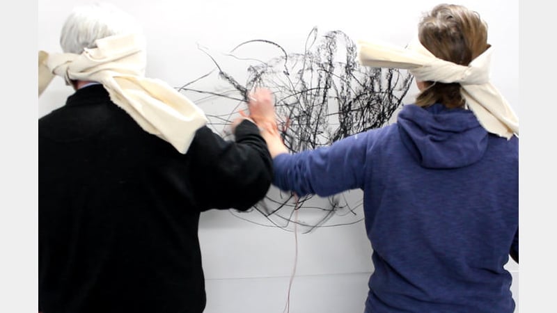 Two students with their backs to the camera draw on a large sheet of paper while blindfolded