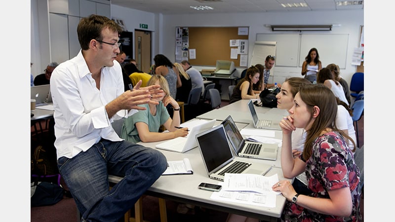 A tutor speaking to a group of students who are sat at a desk with laptops open in front of them